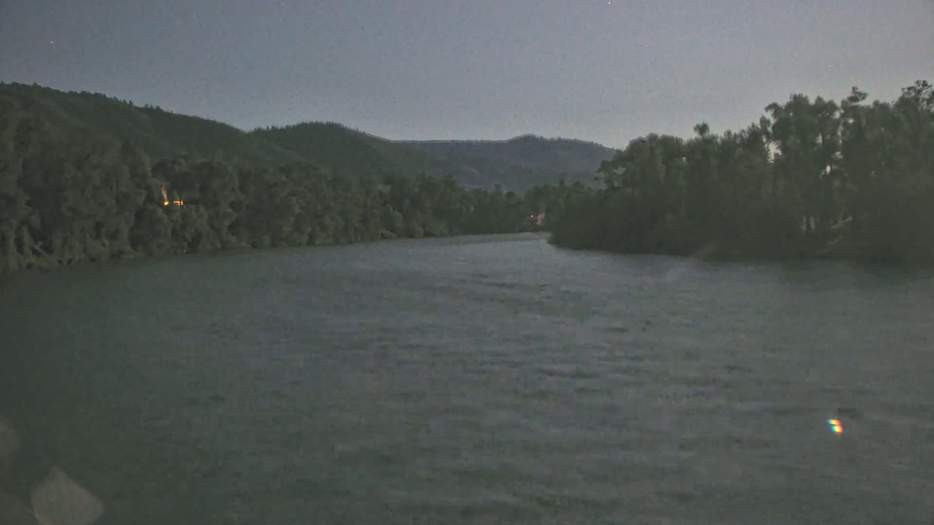 A wide, dark river flows under a twilight sky past tree-lined banks and hills.