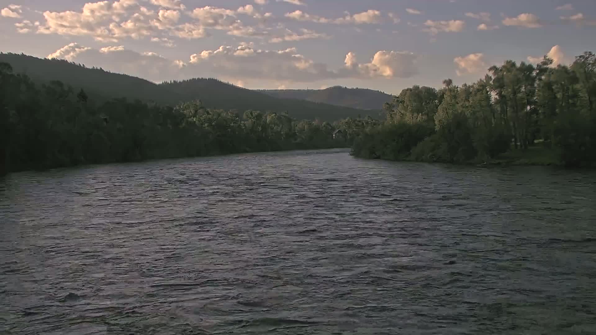 A wide, gently flowing river winds through a valley of dark green trees and hills under a partly cloudy sky.