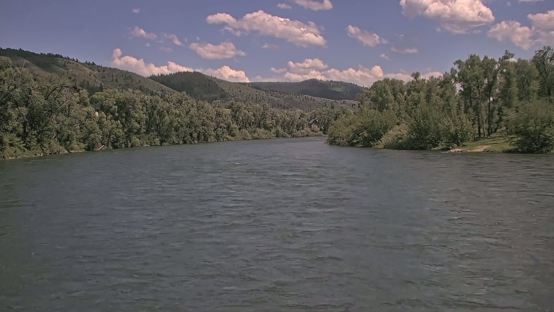 A wide, dark river flows under a partly cloudy sky past lush green banks and hills.