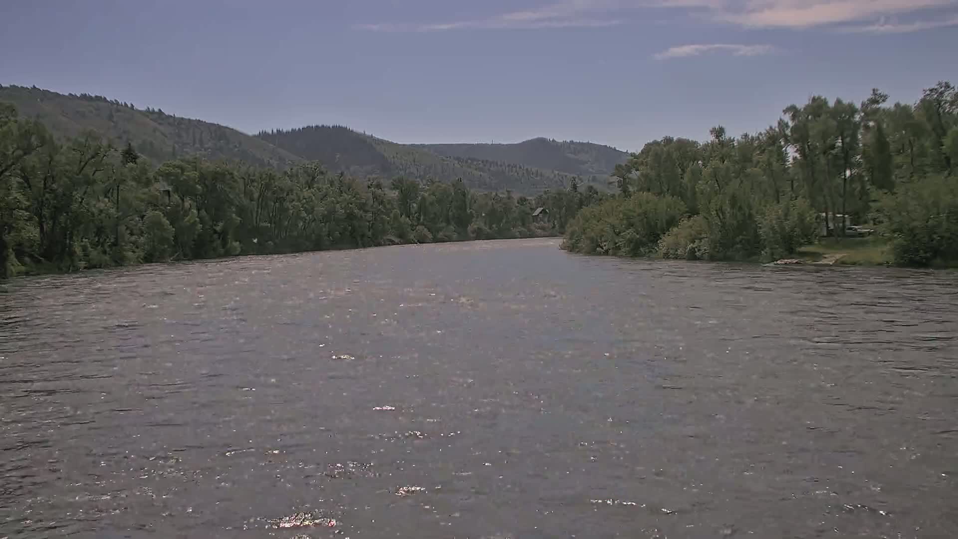 A wide, gently flowing river is seen under a mostly clear sky, bordered by lush green trees and distant hills.