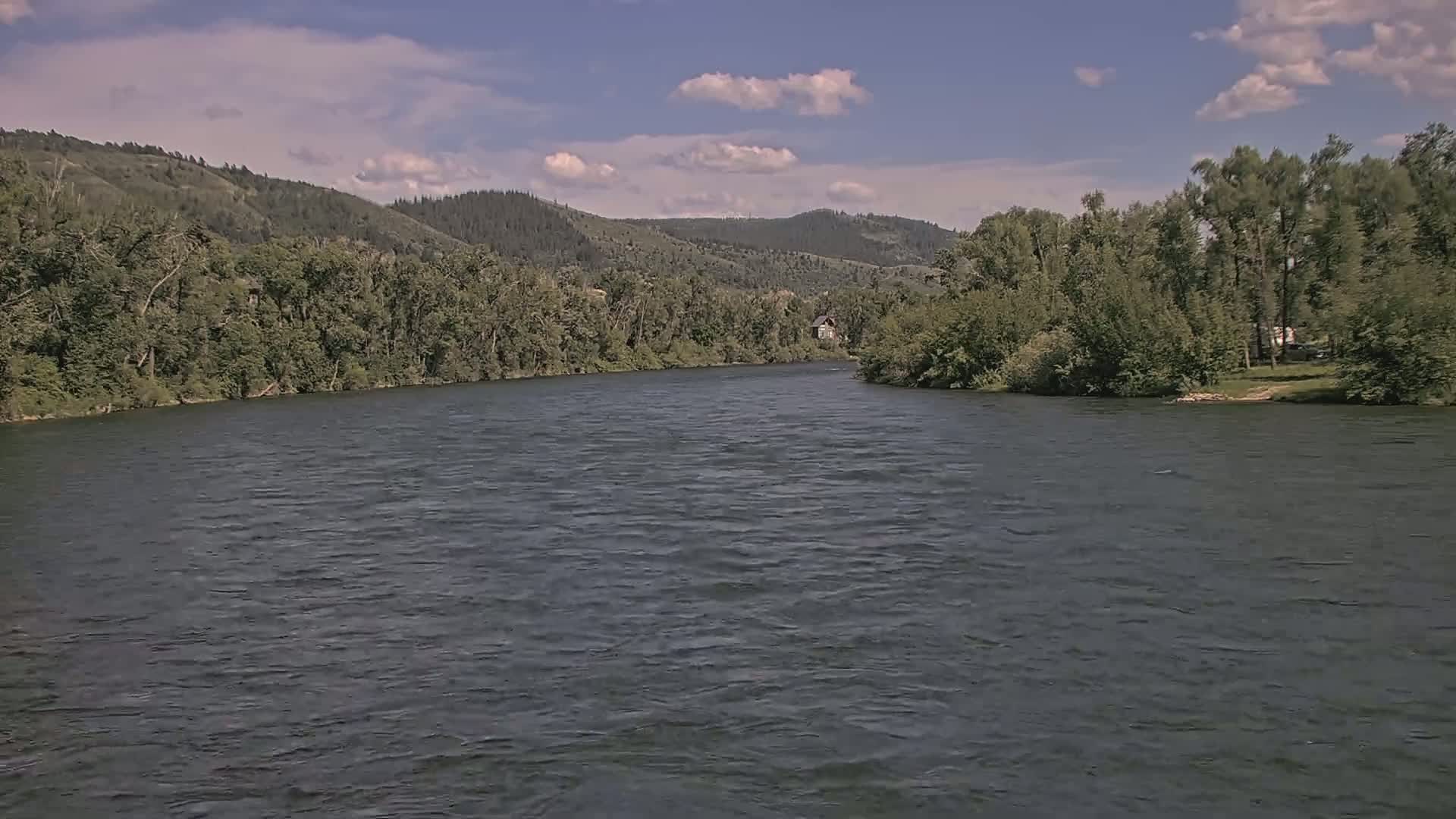 A wide, gently flowing river is seen under a mostly clear sky, bordered by lush green trees and distant hills.