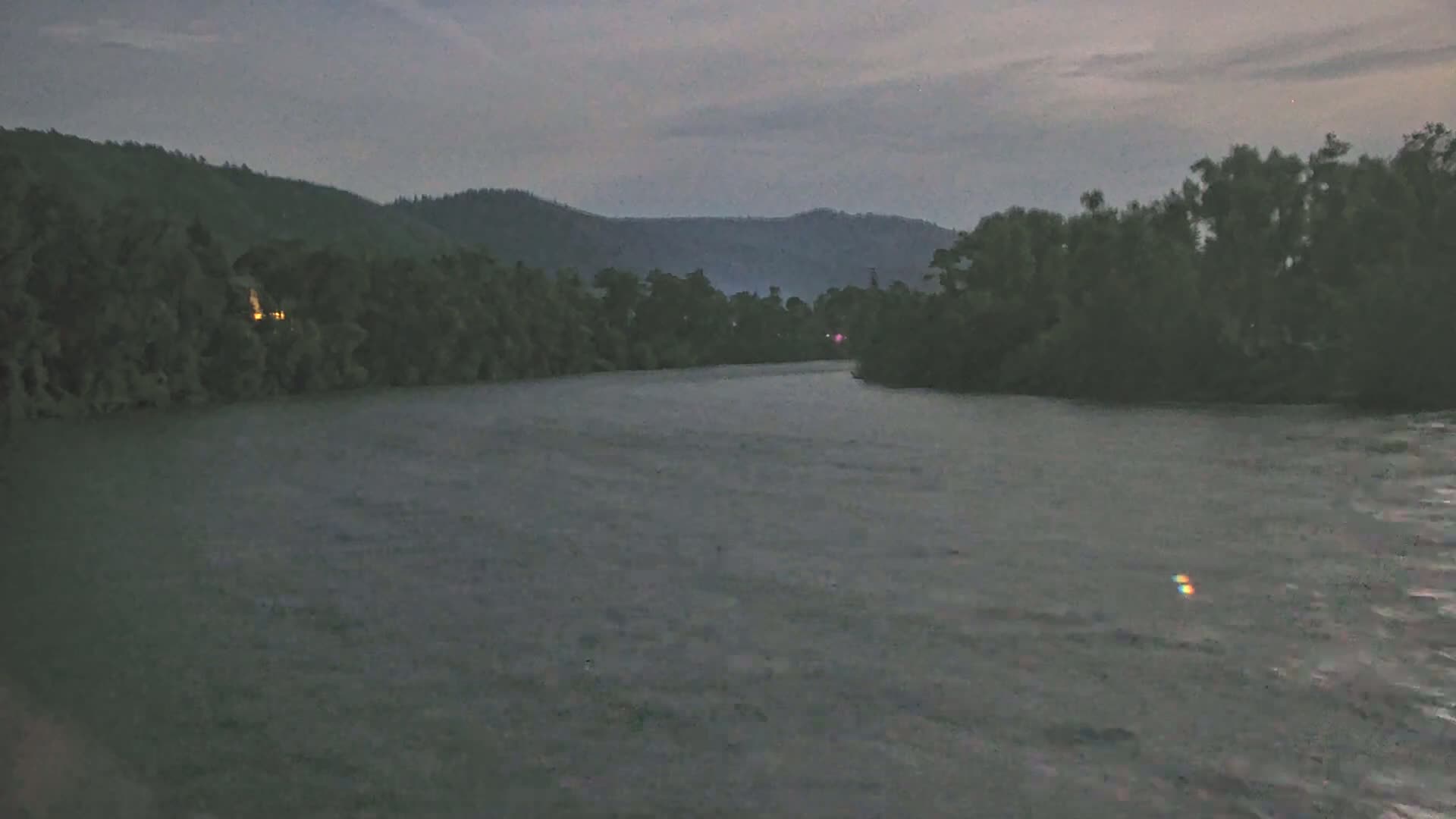 A wide, dark river flows between banks lined with dense trees under a dusky, overcast sky.