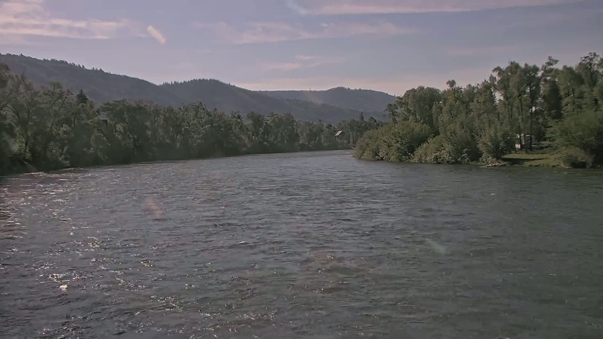 A wide, gently flowing river is surrounded by lush green trees and hills under a mostly clear sky.