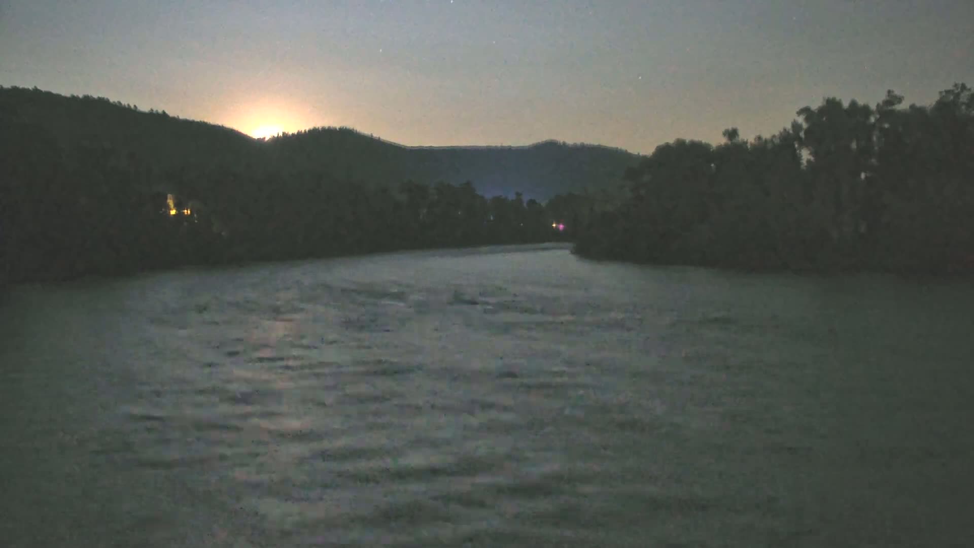 A moonlit night shows a dark, gently rippling river flowing past silhouetted trees and hills under a clear sky.