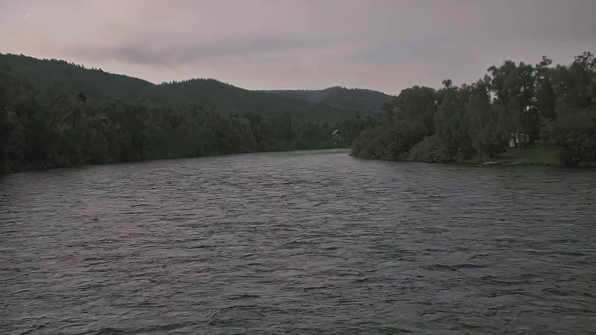 A wide, dark river flows under an overcast sky past a line of dark green trees and hills.