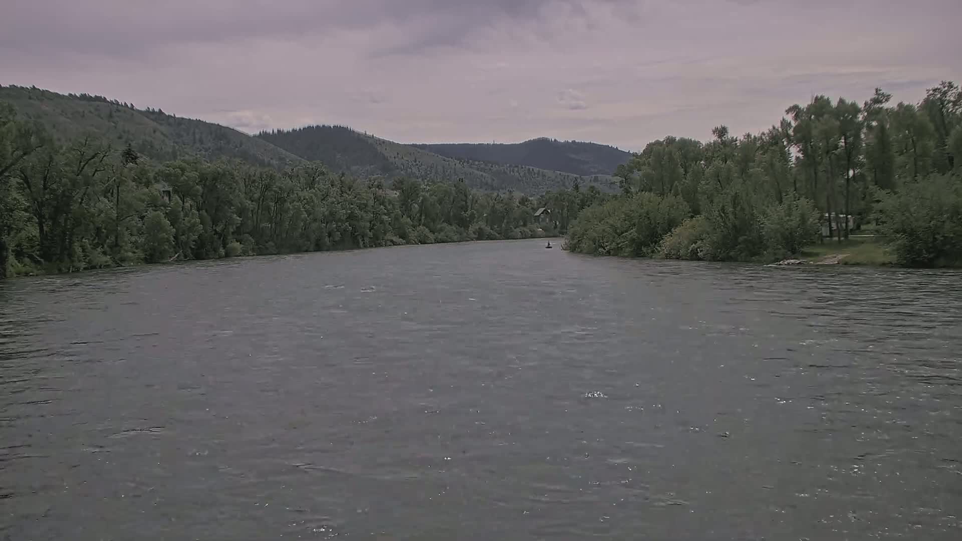 A wide, grey river flows under an overcast sky past lush green banks and distant hills.