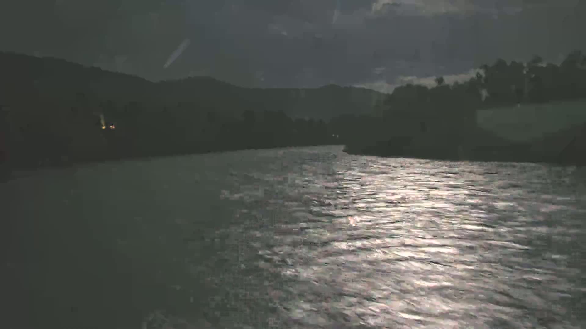A moonlit river flows under a dark, cloudy sky past silhouetted hills and trees.