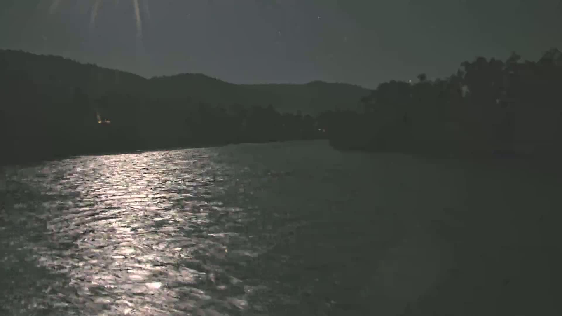 A moonlit river flows under a night sky past dark silhouettes of trees and hills.