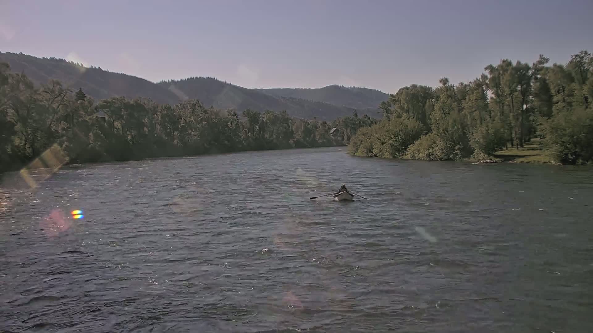 A person rows a small boat on a wide river under a clear sky, with lush green trees lining the banks and mountains in the distance.