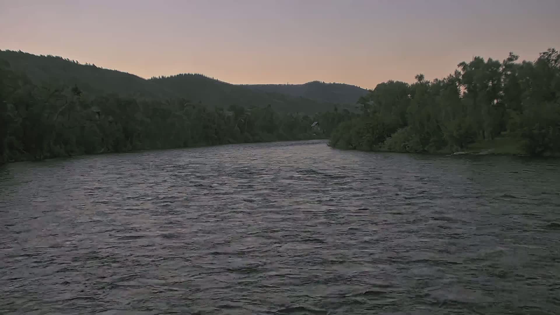 A wide, dark river flows gently under a dusky sky, bordered by lush green hills and trees.