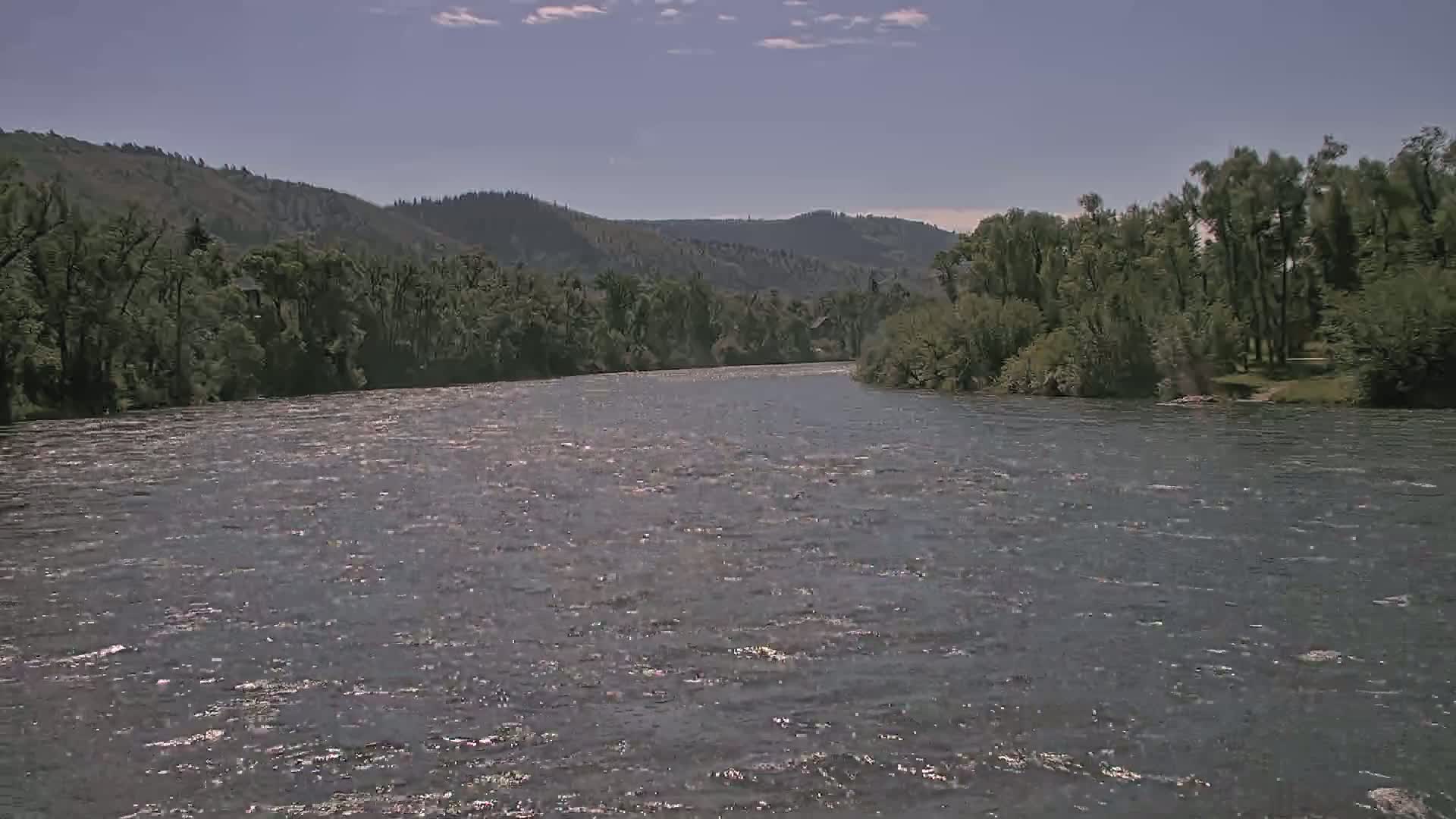 A wide, gently flowing river under a mostly clear sky is bordered by lush green trees and hills.