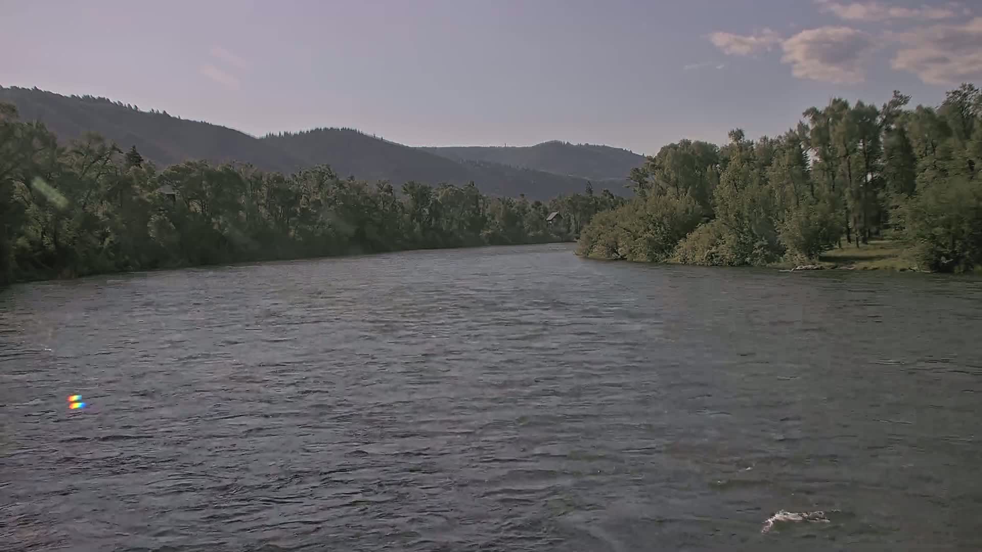 A wide, calm river flows beneath a partly cloudy sky, past lush green banks and forested hills in the background.