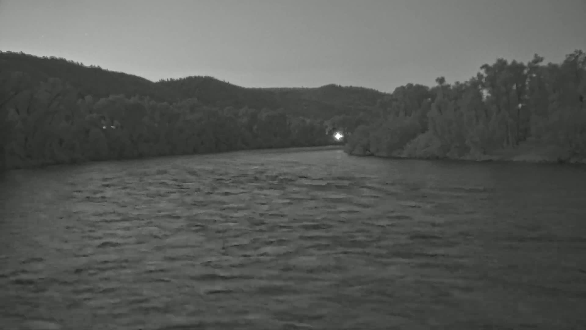 A dark, slightly choppy river flows past a tree-lined bank under a twilight sky.