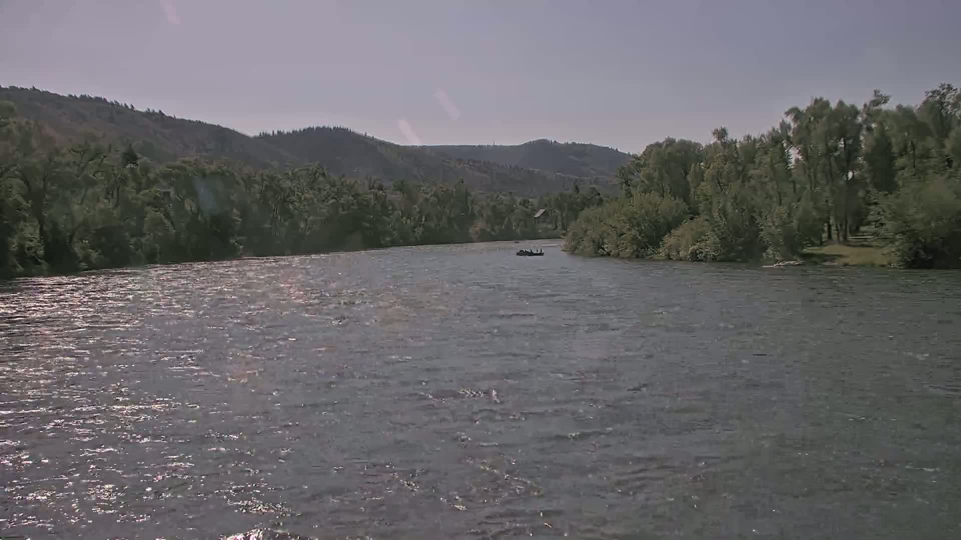 A wide, gently flowing river is seen under a bright, sunny sky, with lush green vegetation and distant hills lining its banks; a small boat is visible in the middle distance.