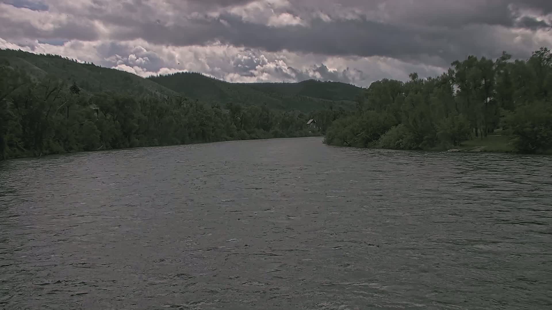 A wide, dark river flows under a cloudy sky toward forested hills.
