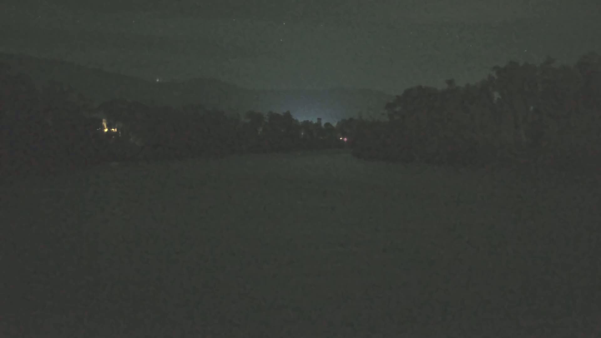 A dark, nighttime scene shows a line of trees and distant lights against a hilly backdrop under a mostly clear sky.