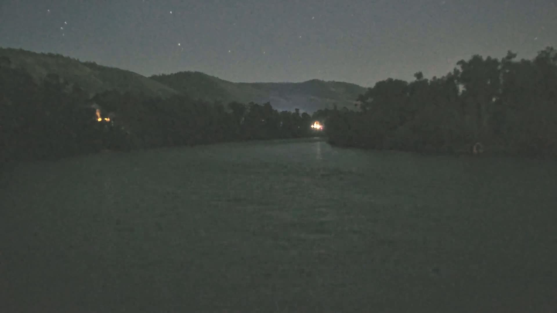 A dark, calm body of water is seen under a clear, starry night sky, with dark, tree-lined banks and hills in the background.