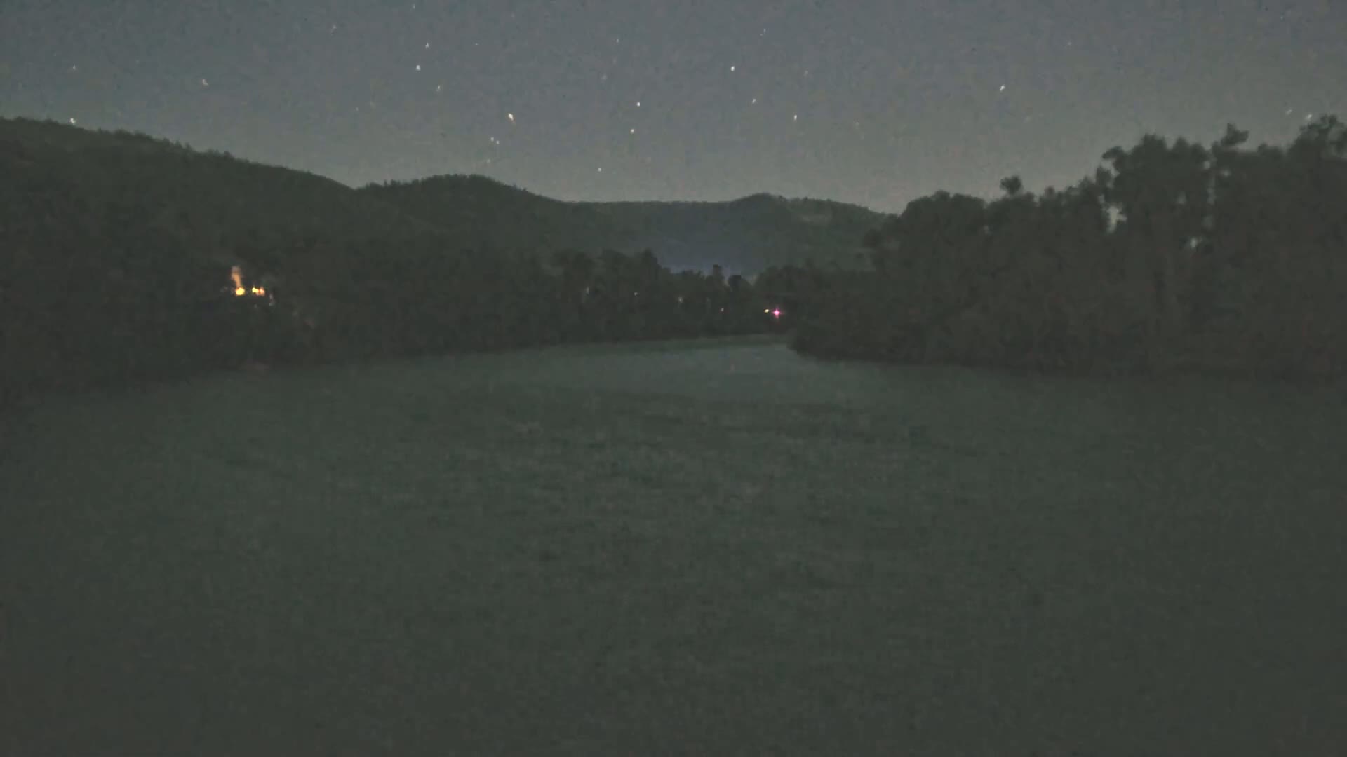 A dark, still body of water is surrounded by trees and hills under a clear, starry night sky.