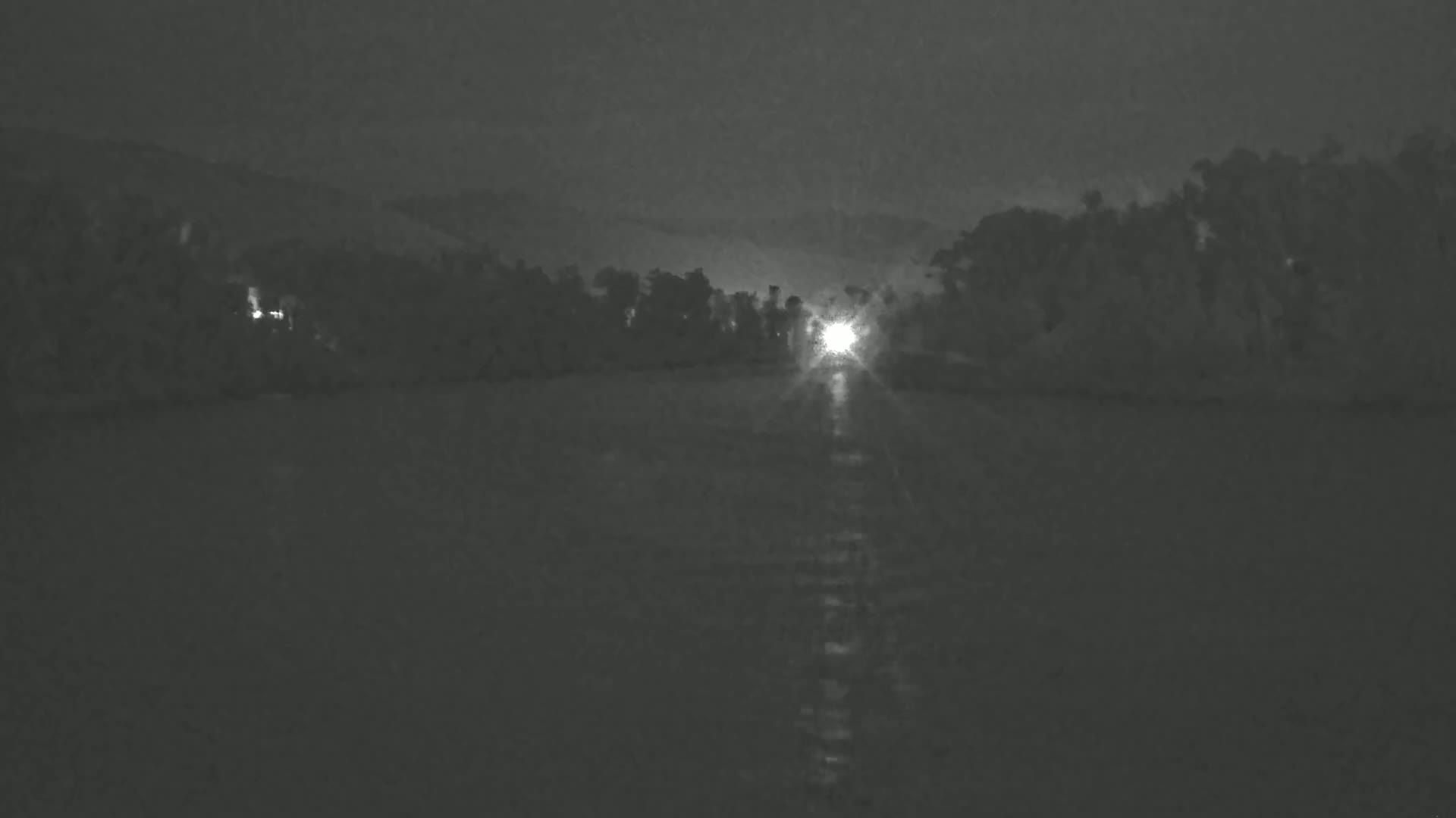 A bright light shines from behind a dark line of trees onto a dark body of water at night.