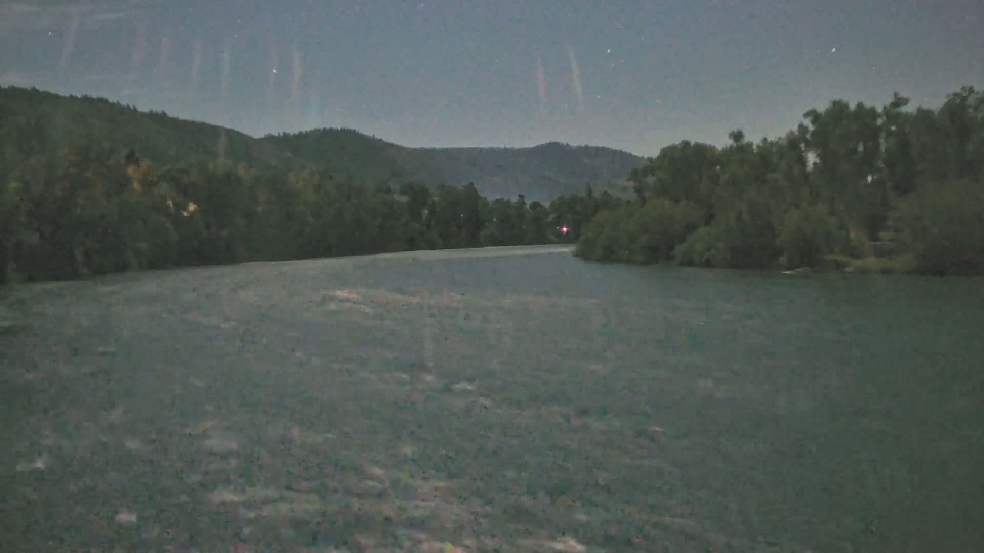 A bright light shines on a dark body of water at night, reflecting a path toward a treeline under a starry sky.
