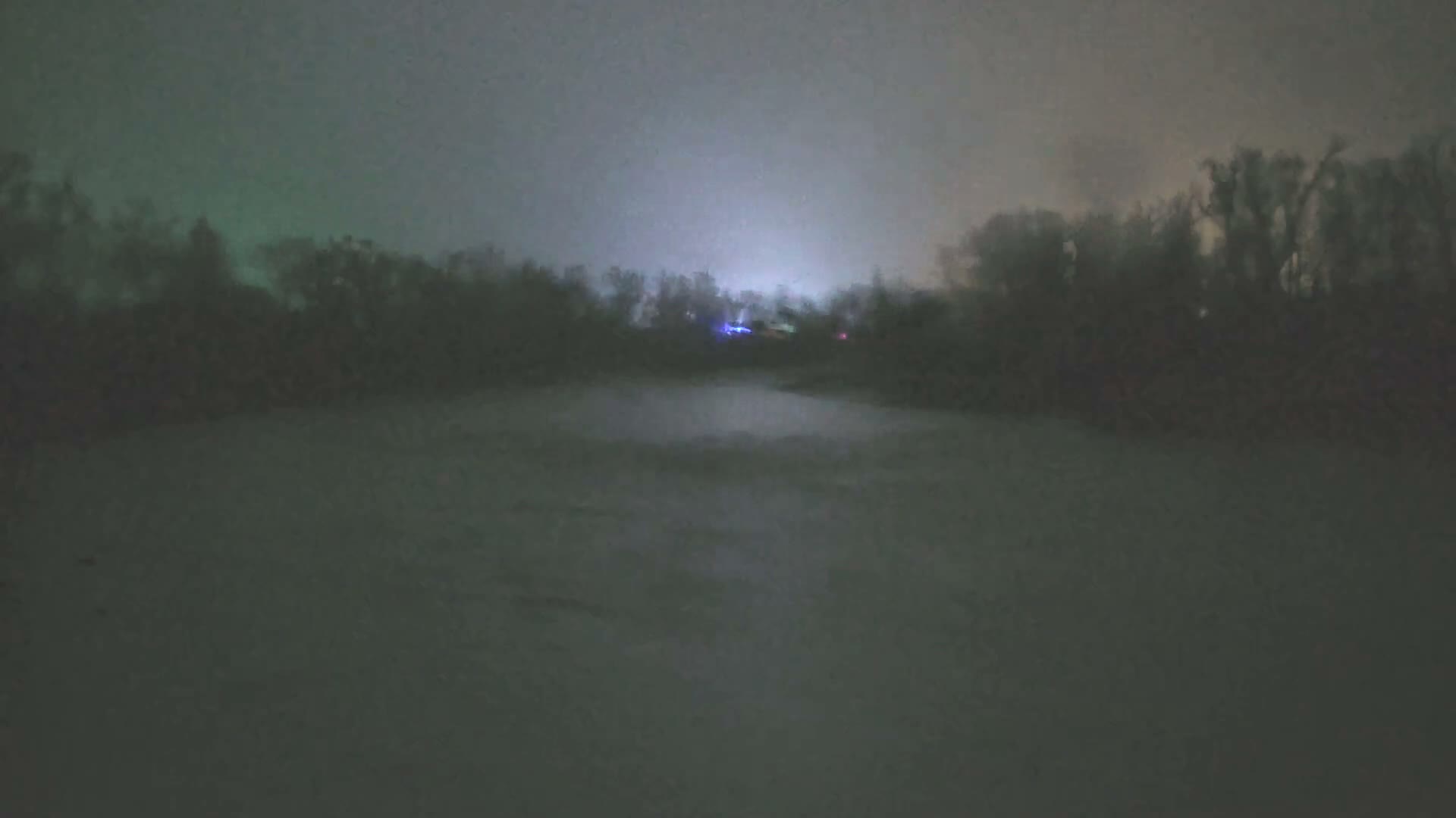A blurry, low-light outdoor image at night shows a dark, flat foreground leading to a line of silhouetted trees against a hazy, greenish-gray sky with a bright, distant light source.