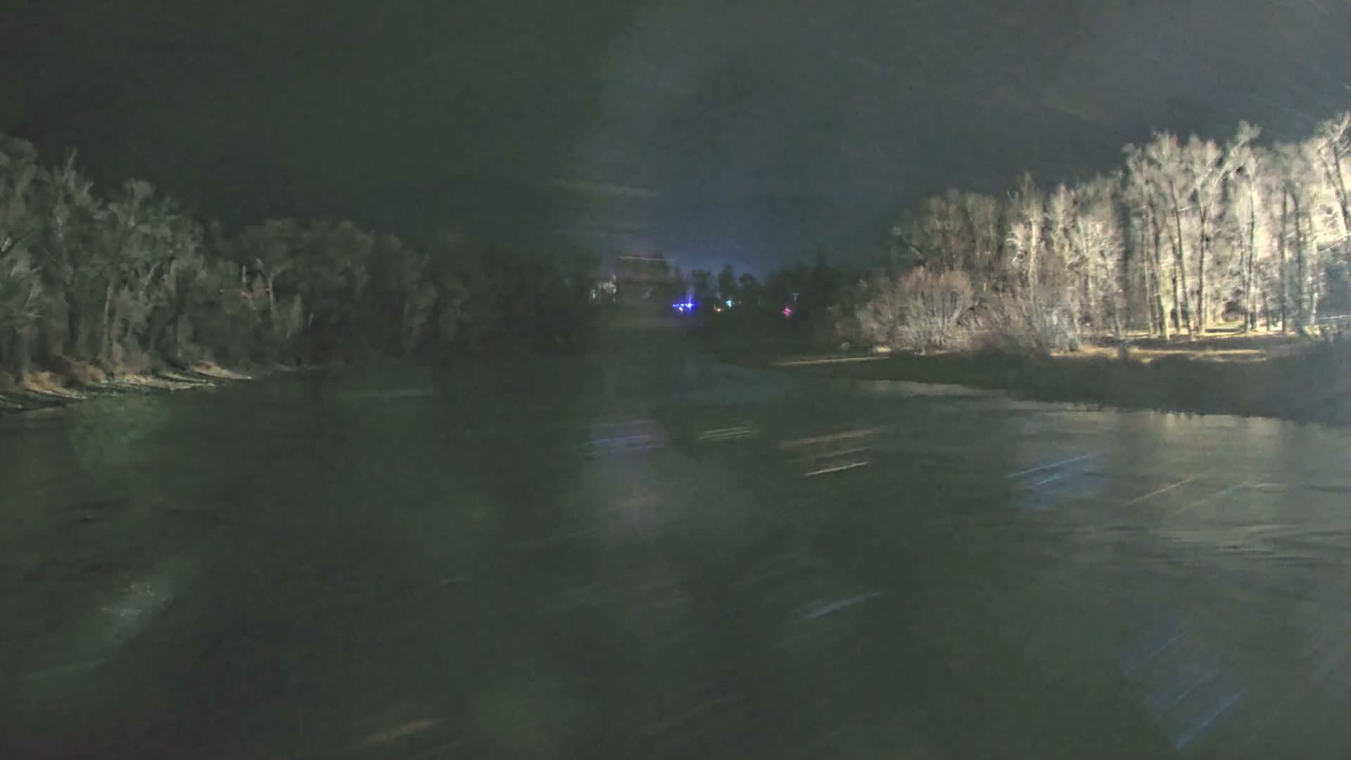 A dark, blurry nighttime scene reveals a wide body of water flanked by dense trees on the left and lighter, bare trees on the right, with faint colorful distant lights reflecting on the water's surface under a dark sky.
