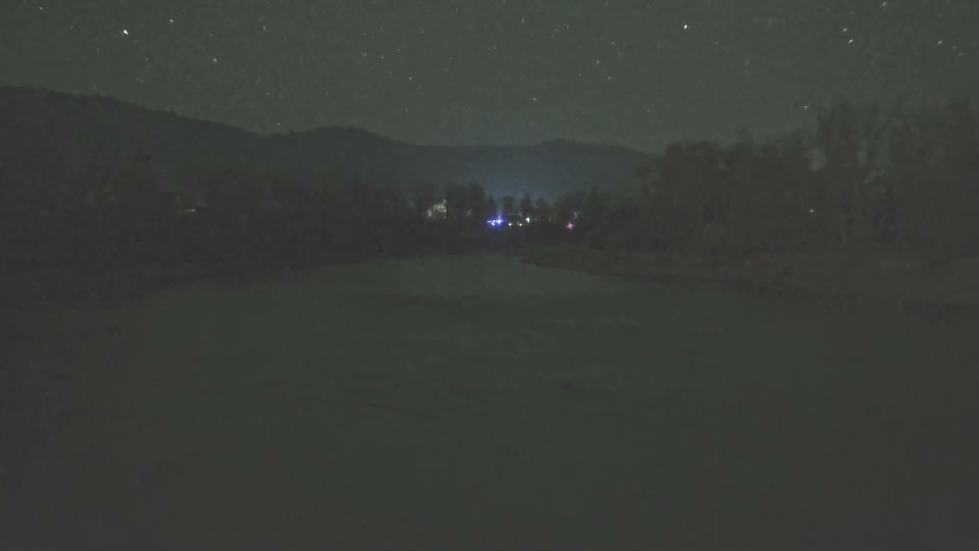 A wide-angle, low-light shot reveals a clear starry night sky above dark hills, with a calm body of water or road in the foreground leading towards distant blue and white artificial lights.