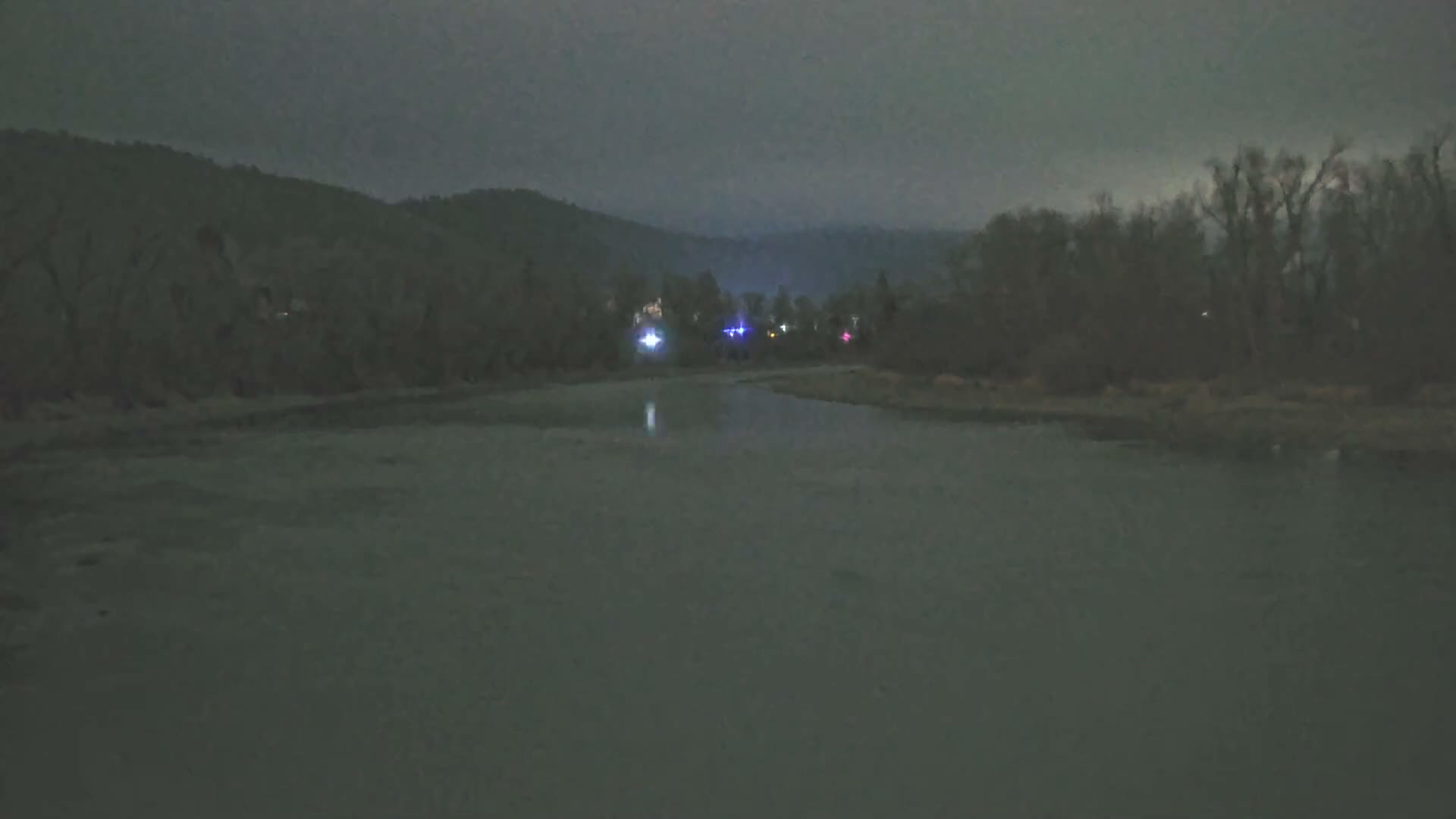 A wide river flows between dark, tree-covered hills under a cloudy night sky, with colorful artificial lights reflecting on the far bank.