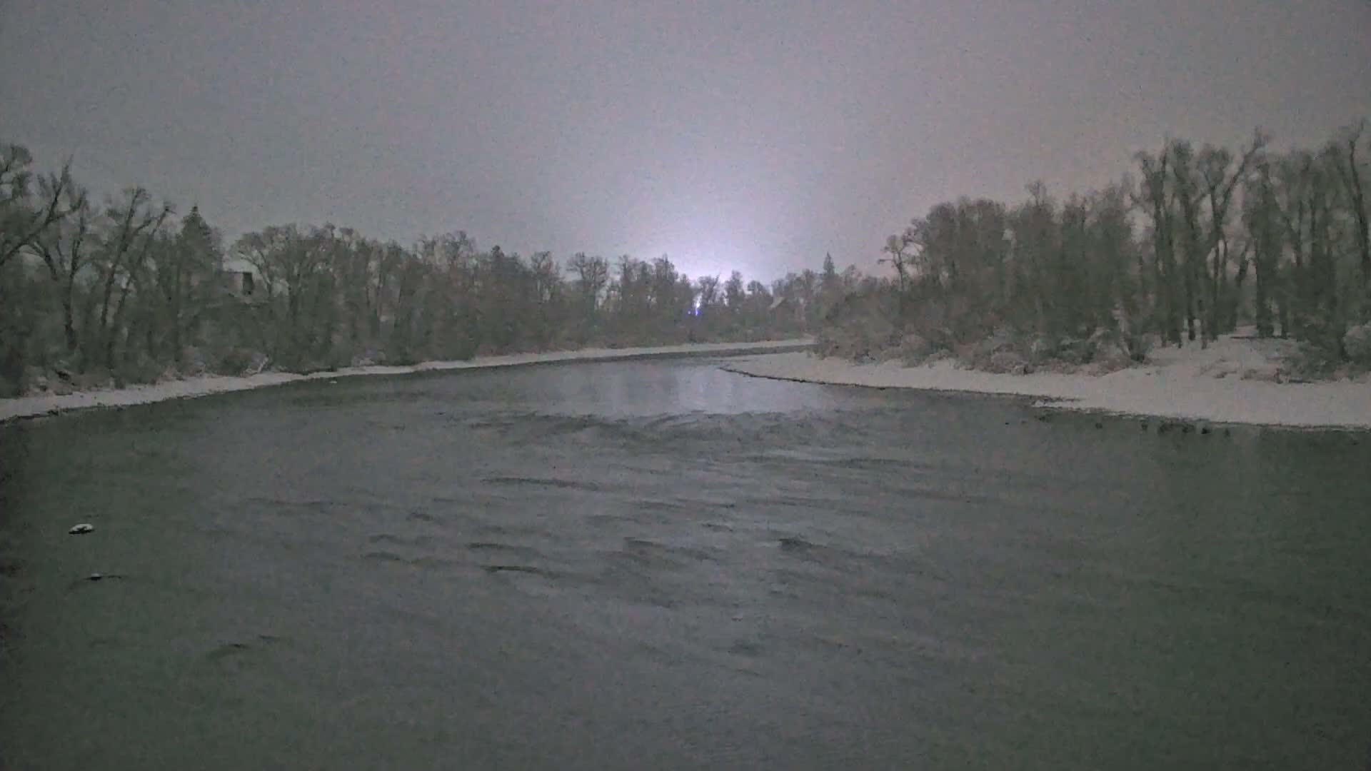 A snow-covered river flows under an overcast, grey sky, flanked by wintry trees and snow-dusted banks, with a bright light visible on the distant horizon during what appears to be a snowy, cold day or evening.