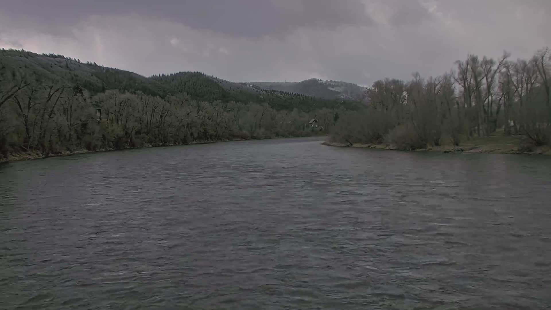 A snow-covered river flows under an overcast, grey sky, flanked by wintry trees and snow-dusted banks, with a bright light visible on the distant horizon during what appears to be a snowy, cold day or evening.