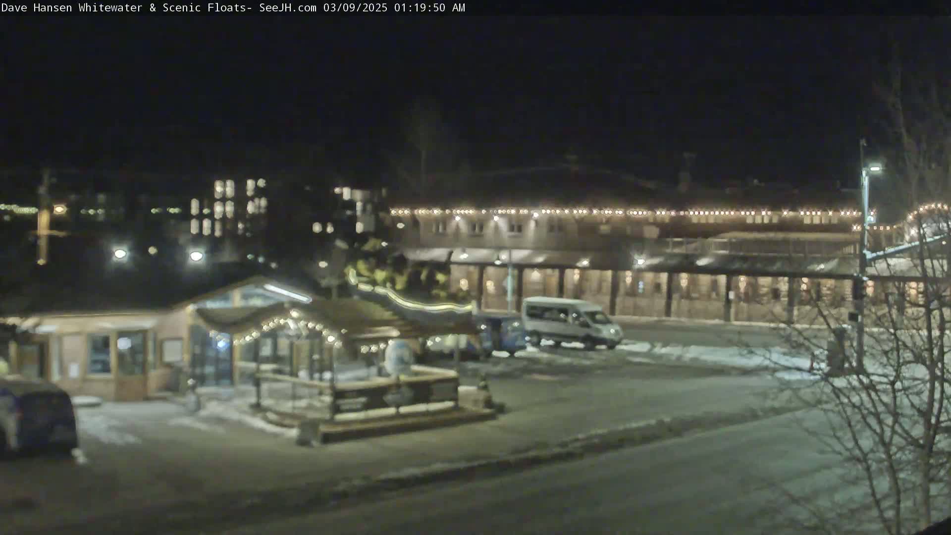A nighttime view of a snow-covered parking lot with several buildings and vehicles, including a van, under partly cloudy skies.