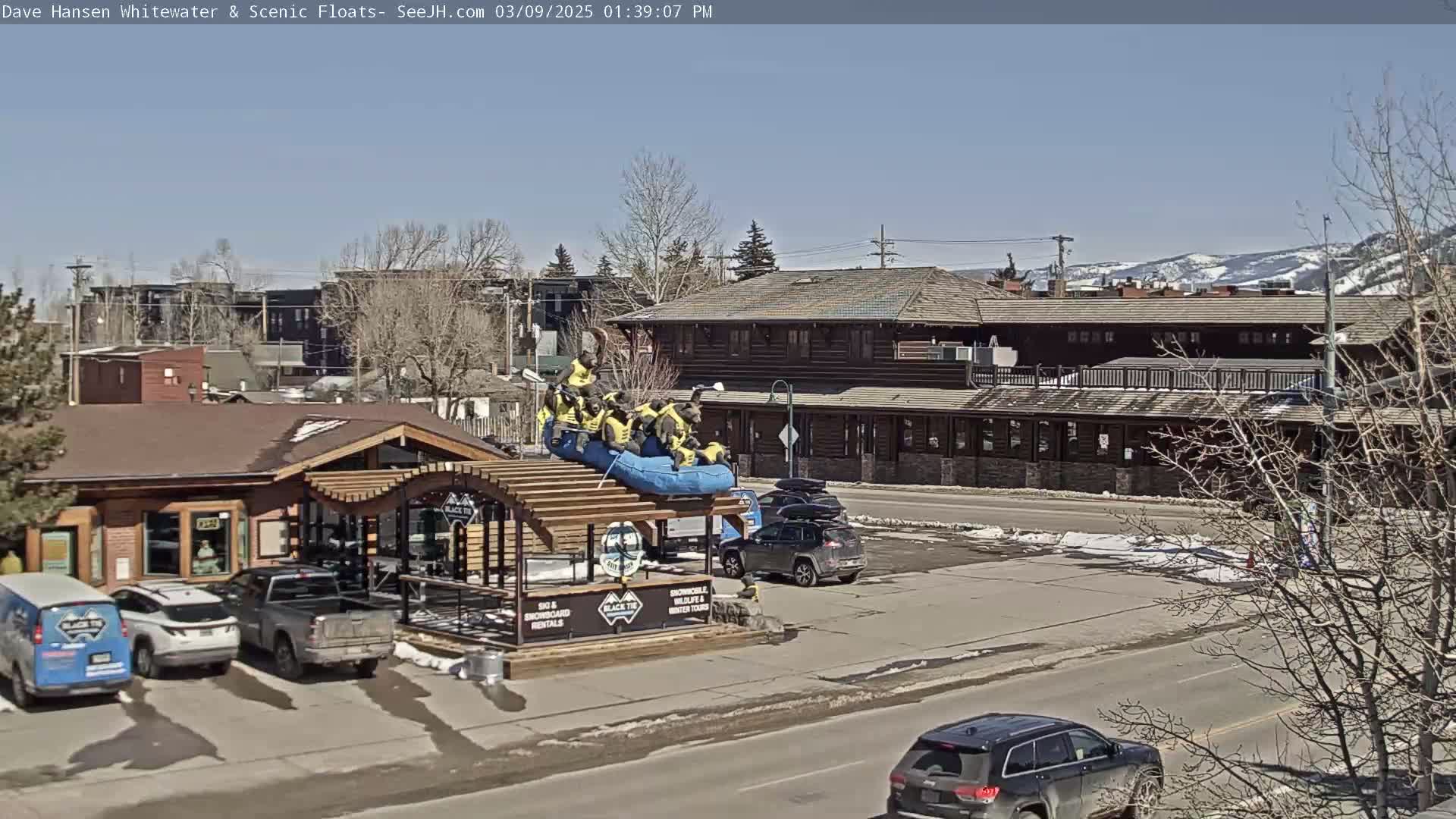 Under a clear blue sky, a group of people in yellow life vests are positioned on a blue raft atop a building, near parked cars and a larger log cabin-style building.
