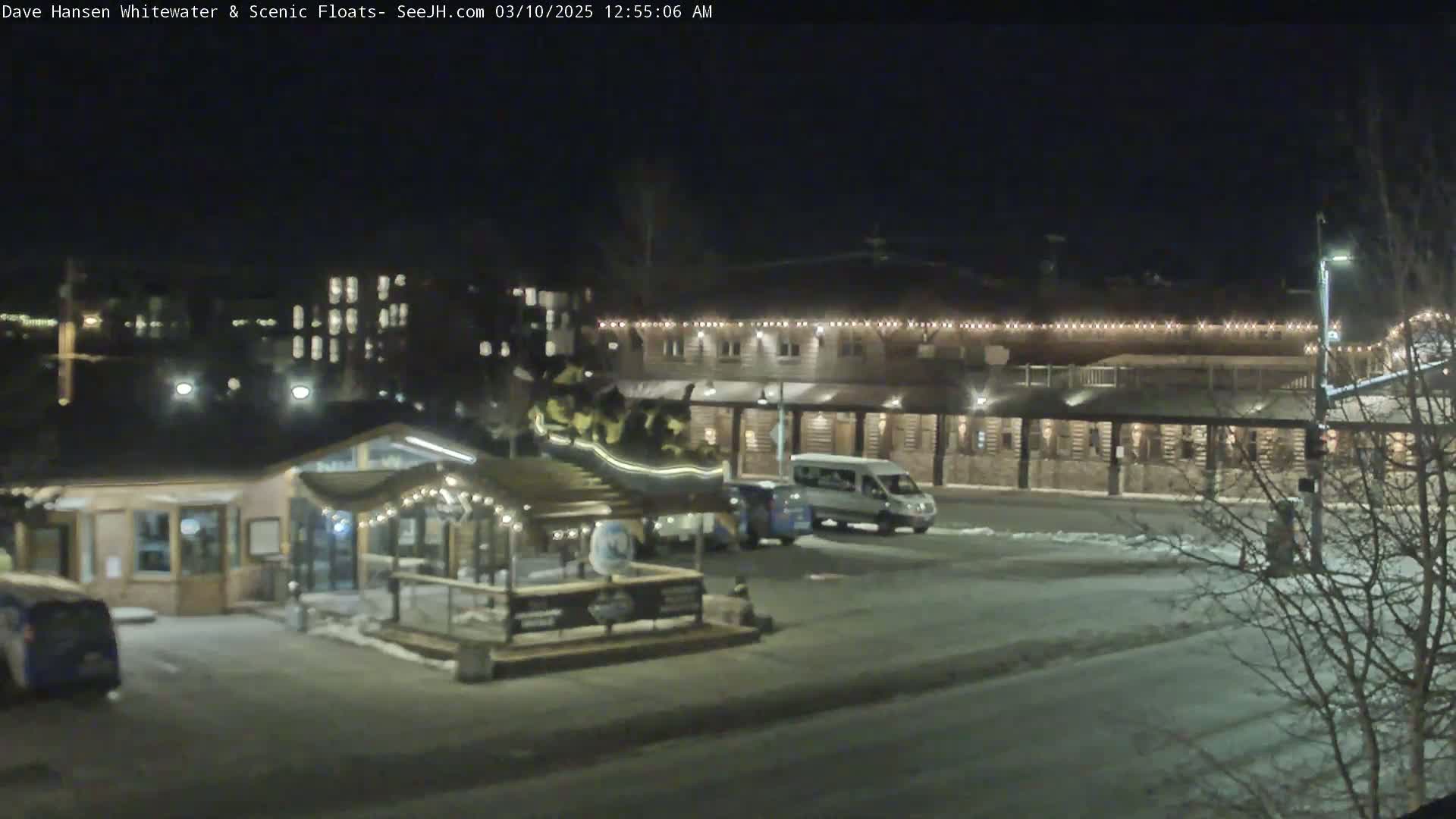 A nighttime view of a snow-covered parking lot with a small building and a larger building with string lights across its facade.