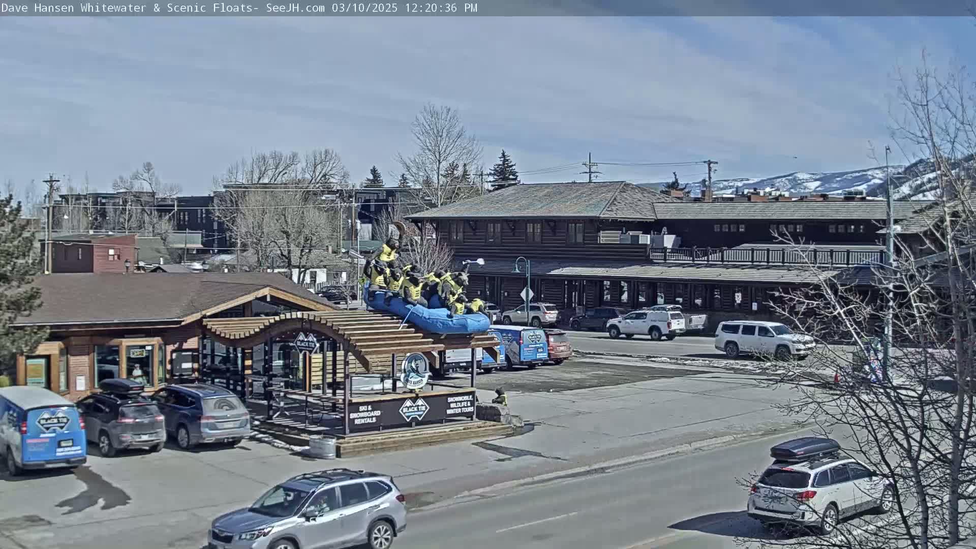 A sunny day shows a group of people in yellow life jackets on a raft atop a building near parked cars and a street.