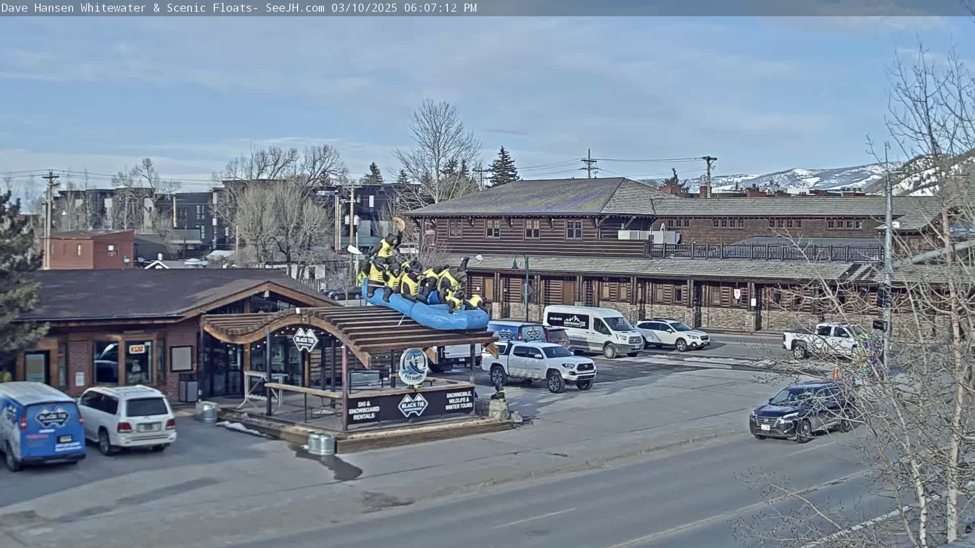 A sunny day reveals a parking lot with several vehicles, a building with a rafting display on its roof, and other buildings in the background, including snow-capped mountains.