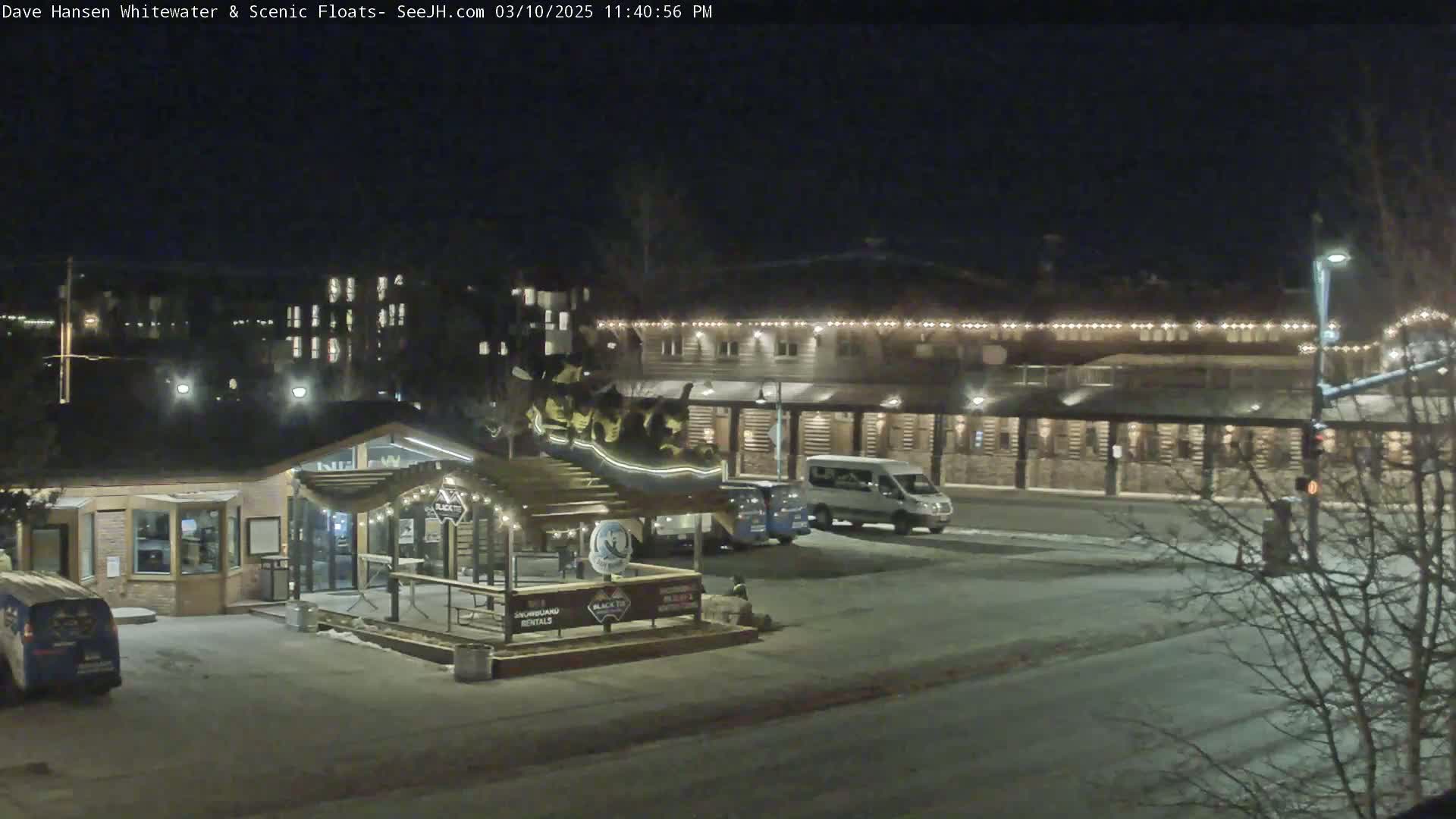 A nighttime view of a building complex with string lights, a few vehicles in the parking lot, and a small covered structure with signage.