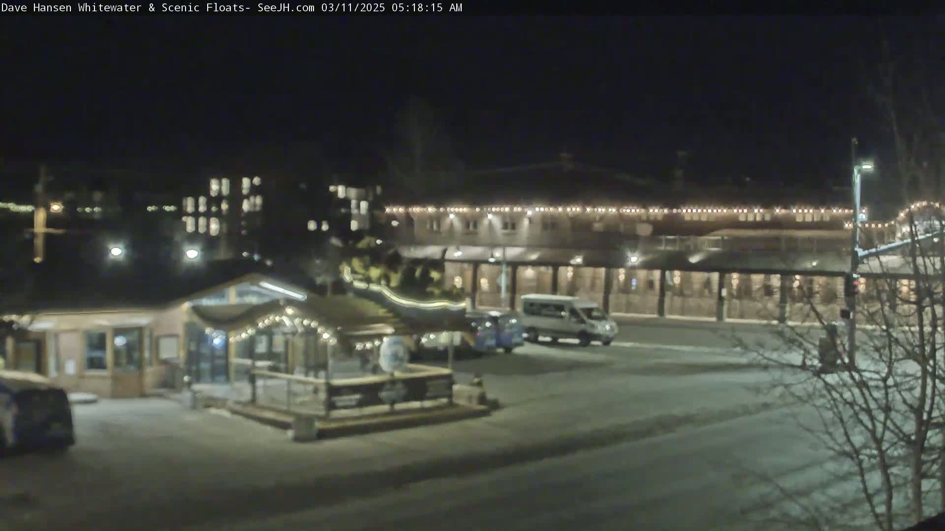 A nighttime, snowy scene shows a parking lot with several vehicles near a small building with string lights and a larger building with illuminated windows in the background.