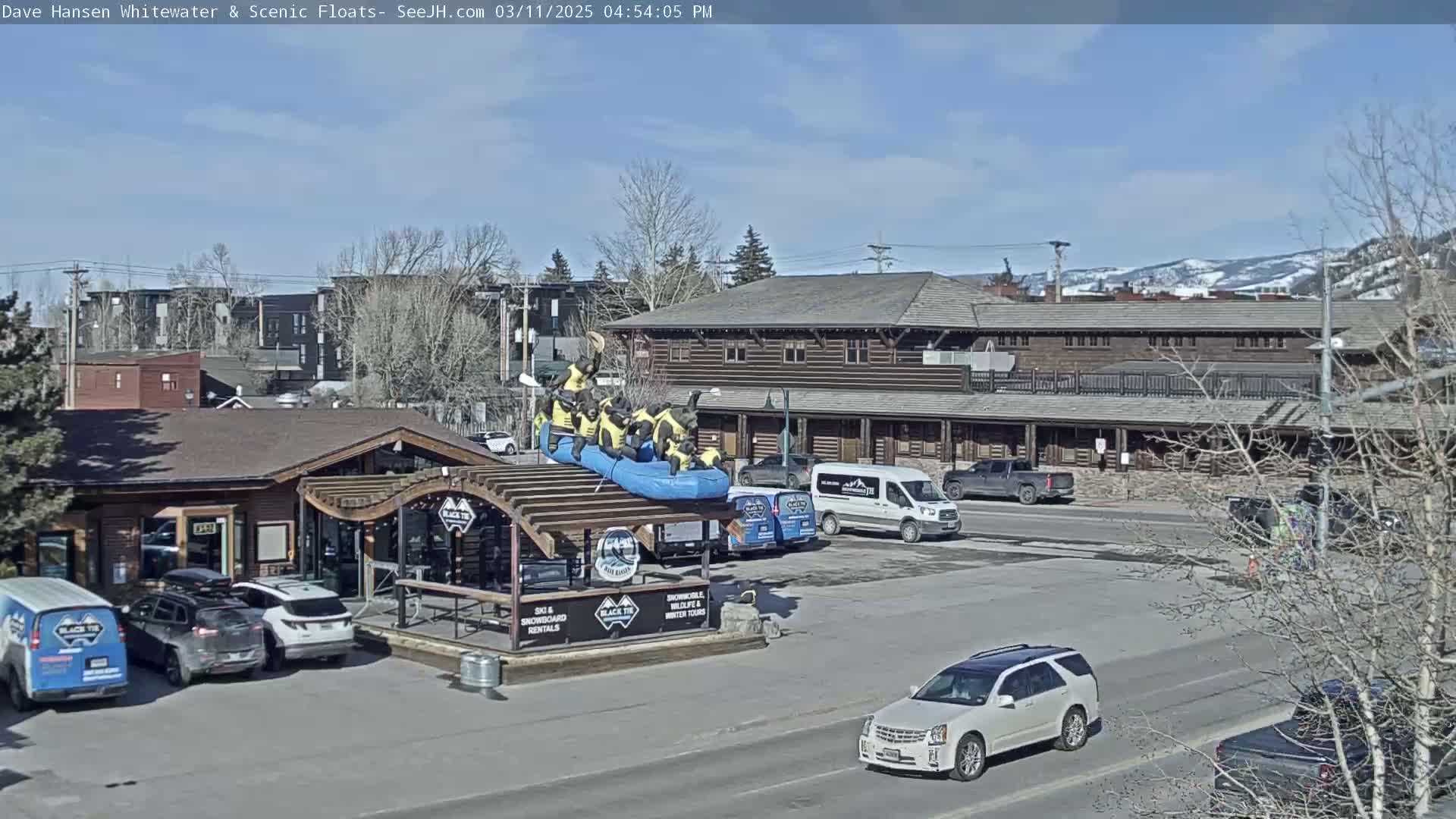 A sunny day shows a parking lot with several vehicles, a building with a rafting display, and a background of mountains and other buildings.