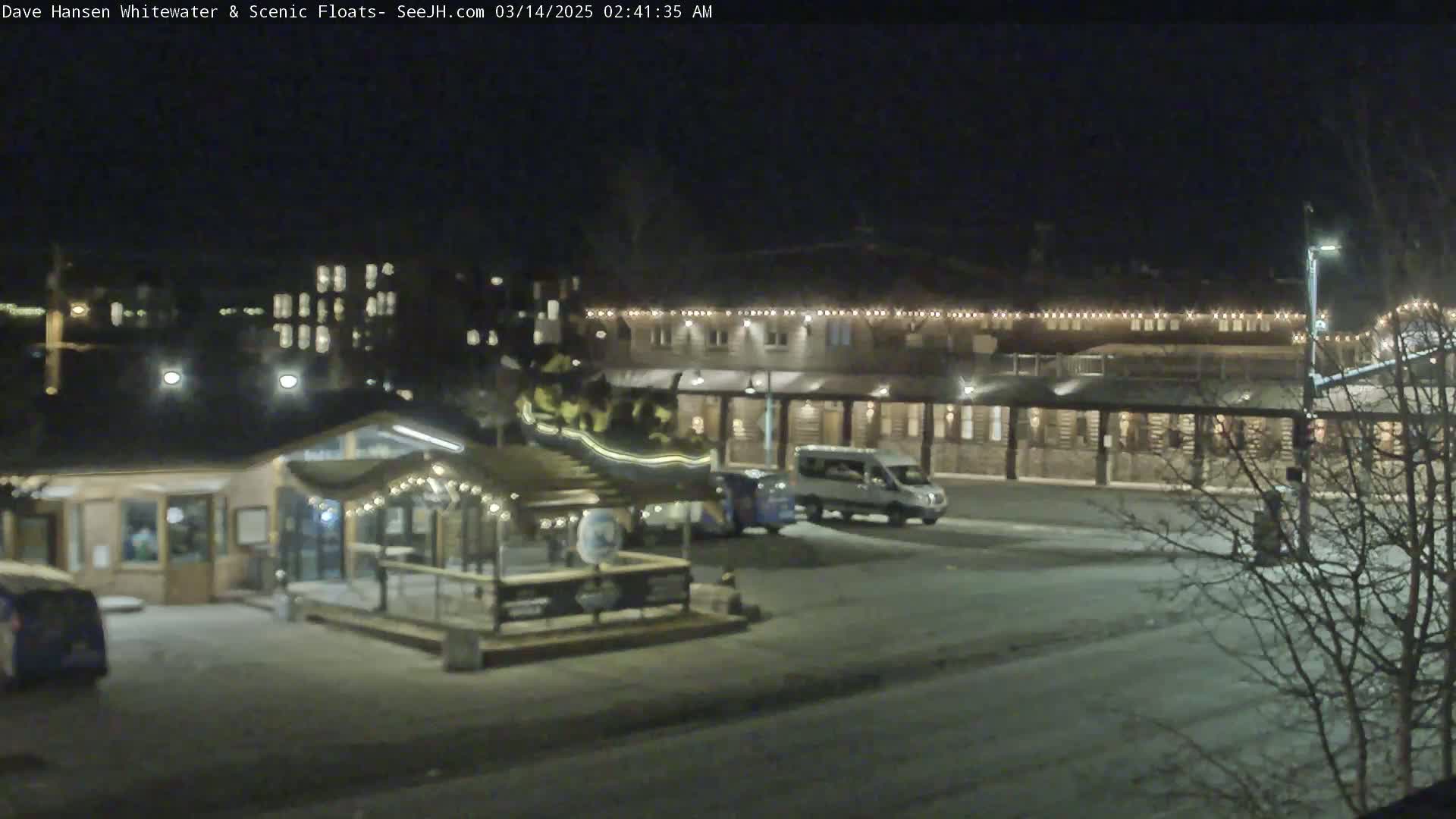 A nighttime view of a snow-covered parking lot in front of a large building with string lights, alongside a smaller building with a covered patio area.