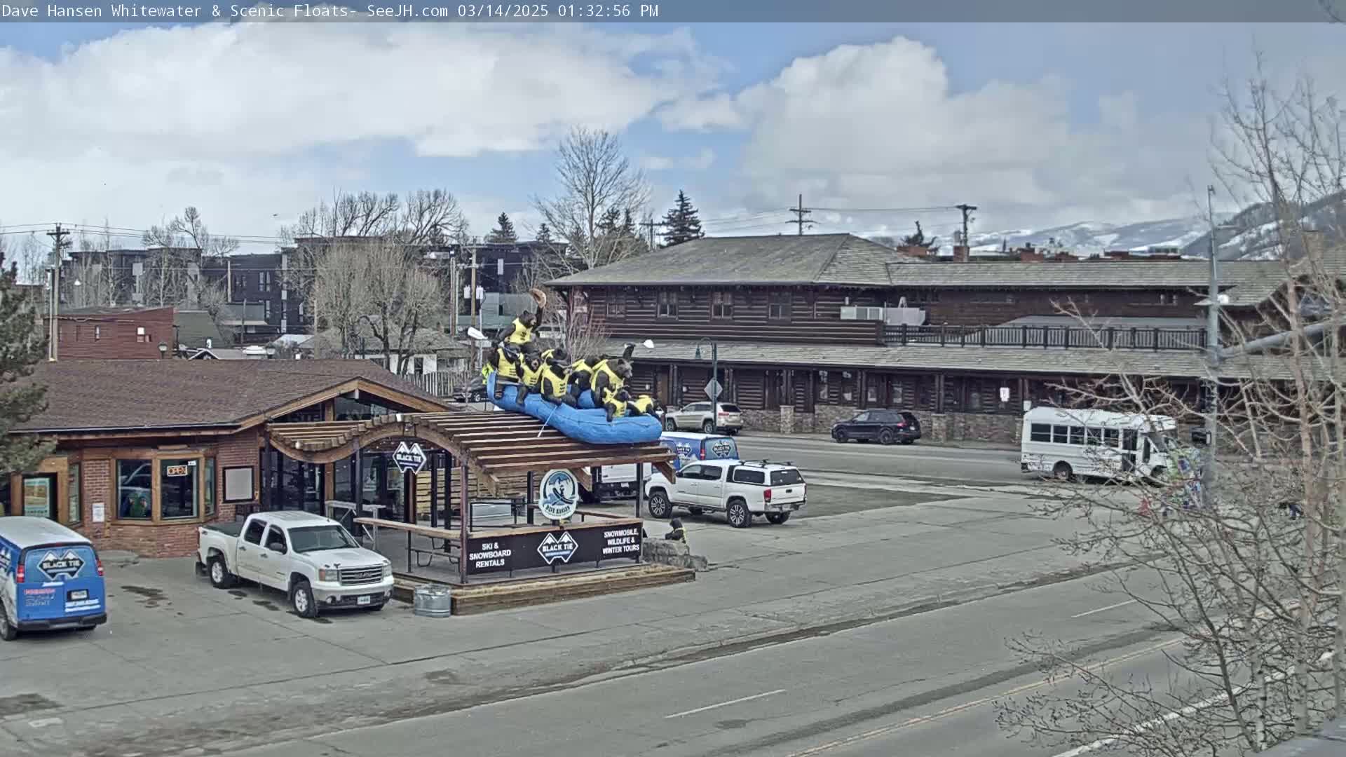 A partly cloudy day shows a log cabin-style building with a rafting-themed display on the roof of an adjacent building, a parking lot with several vehicles including a pickup truck and a van, and a white bus in the background.