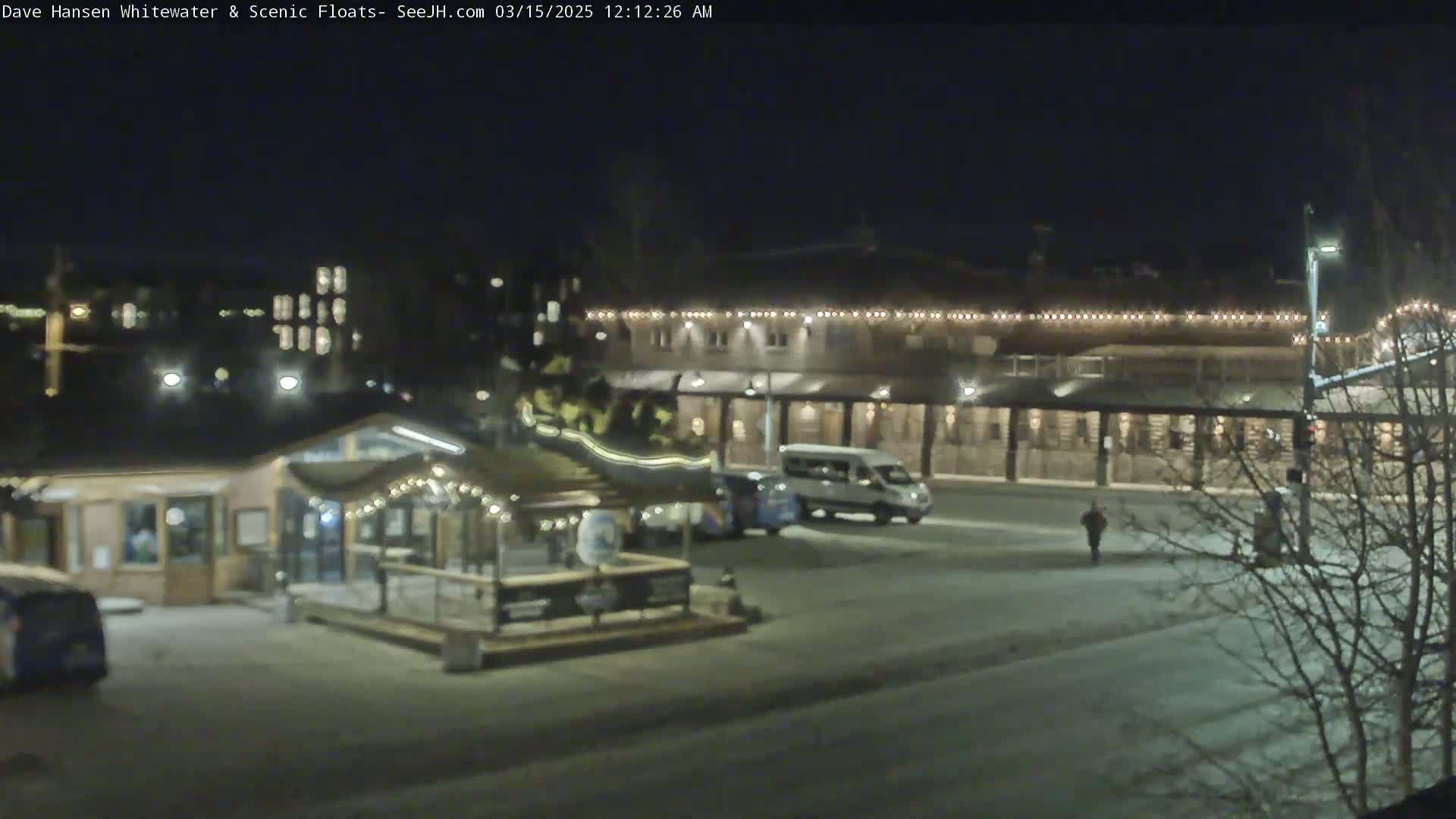 A nighttime view of a parking lot with a small building, a larger building with lights strung along the roofline, and a few vehicles, all covered in a light layer of snow.