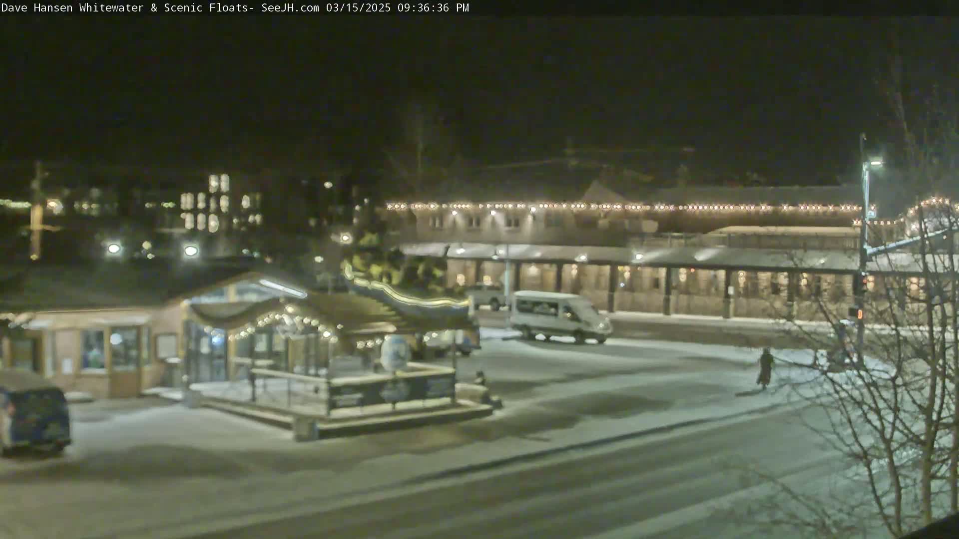 A nighttime scene of a snow-covered town square with buildings, vehicles, and a few pedestrians, illuminated by streetlights.