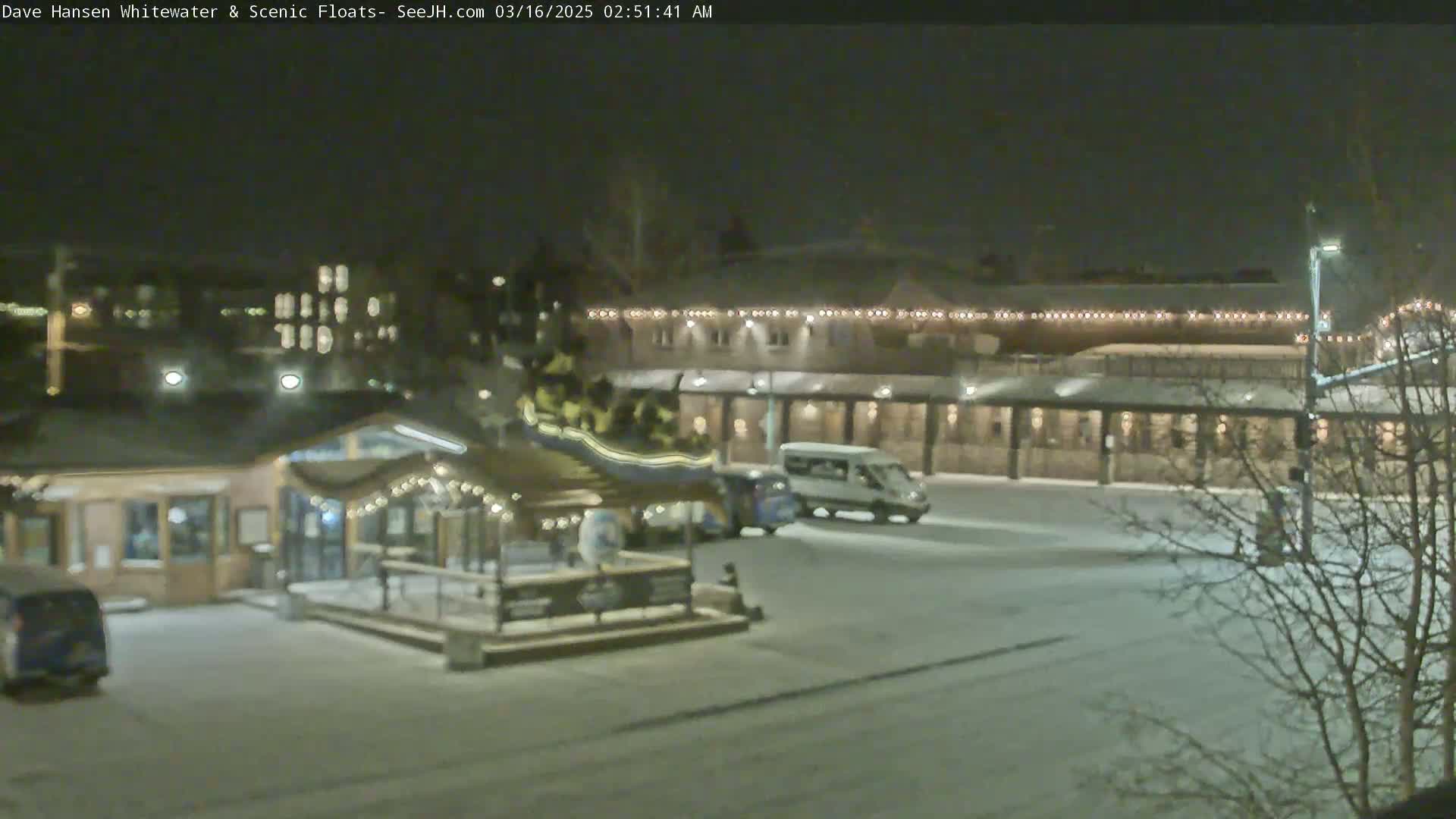 A nighttime, snow-covered parking lot with a small building and a larger building with string lights across the front, and a few vehicles are visible.