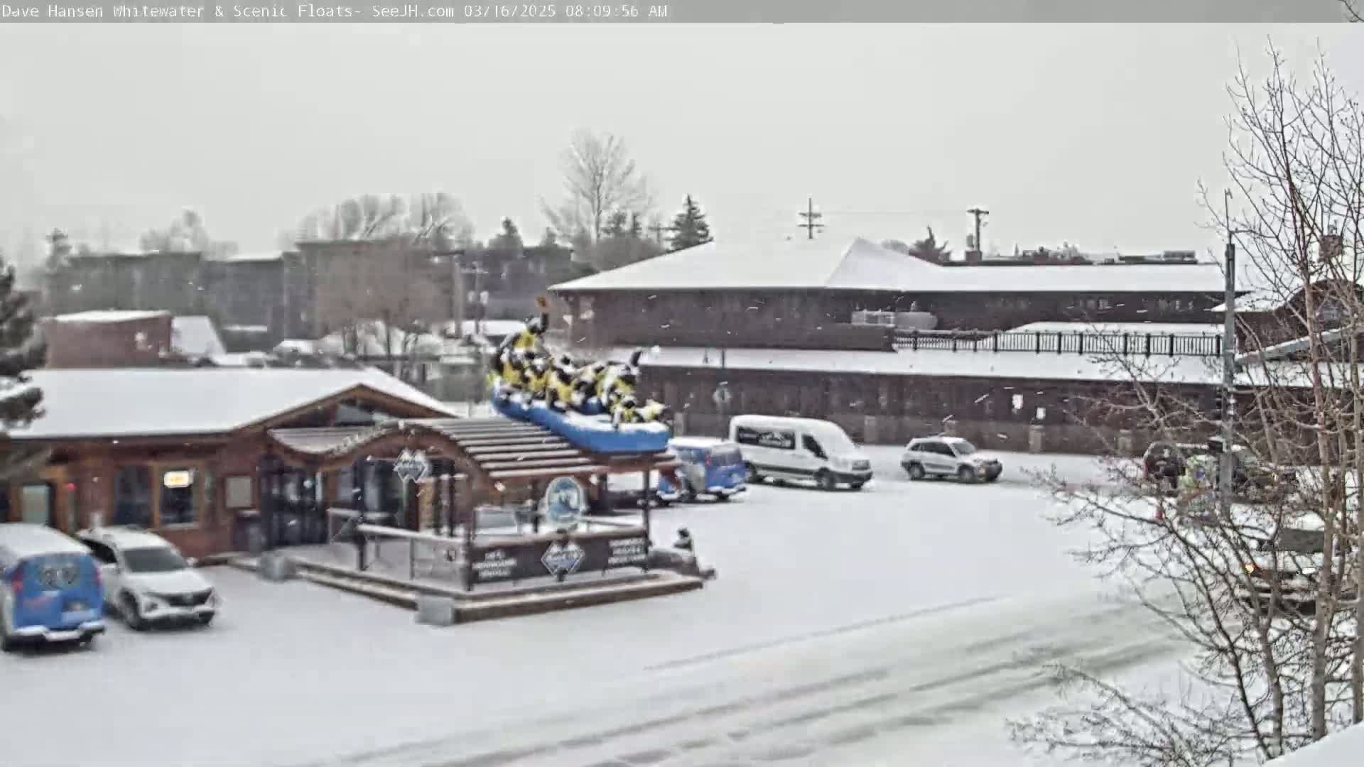 A snow-covered parking lot with several vehicles, a log cabin-style building, and a large inflatable raft on a structure, all under a light snowfall.