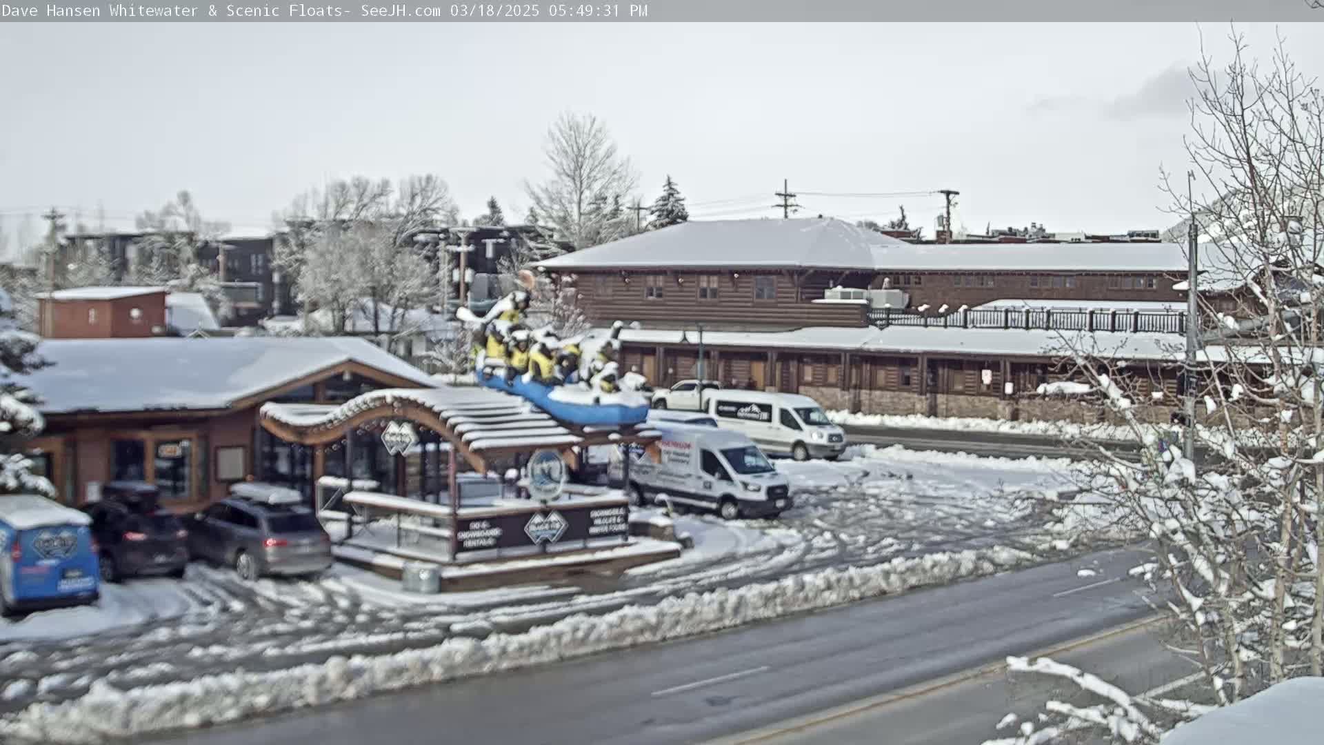 A snow-covered scene shows several buildings, parked cars, and a raft with people on it situated near a snow-covered wooden structure.