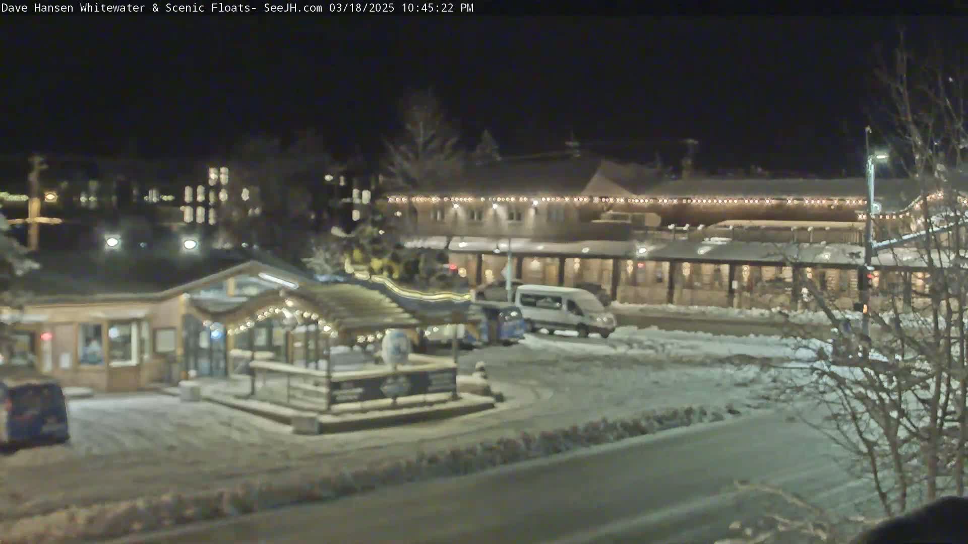 A snow-covered nighttime scene shows a building with string lights, a small structure with a gazebo-like top, and a van parked in a lot.