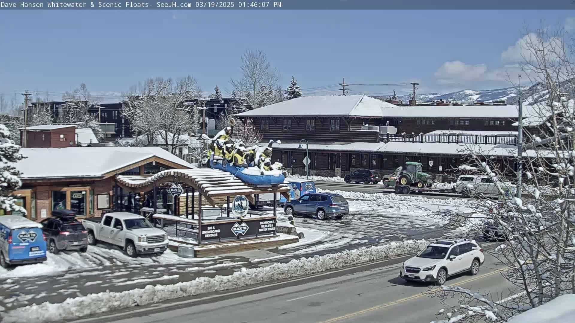 A snow-covered town scene on a sunny day shows a street with parked cars, a building with a raft sculpture in front, and snow-dusted trees.