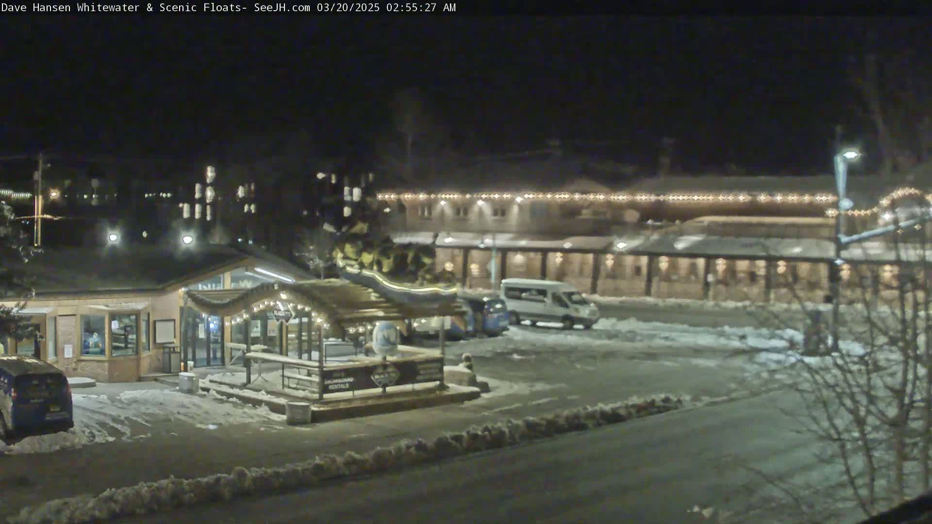 A nighttime view of a snow-covered parking lot with a building illuminated by lights, and several vehicles parked.