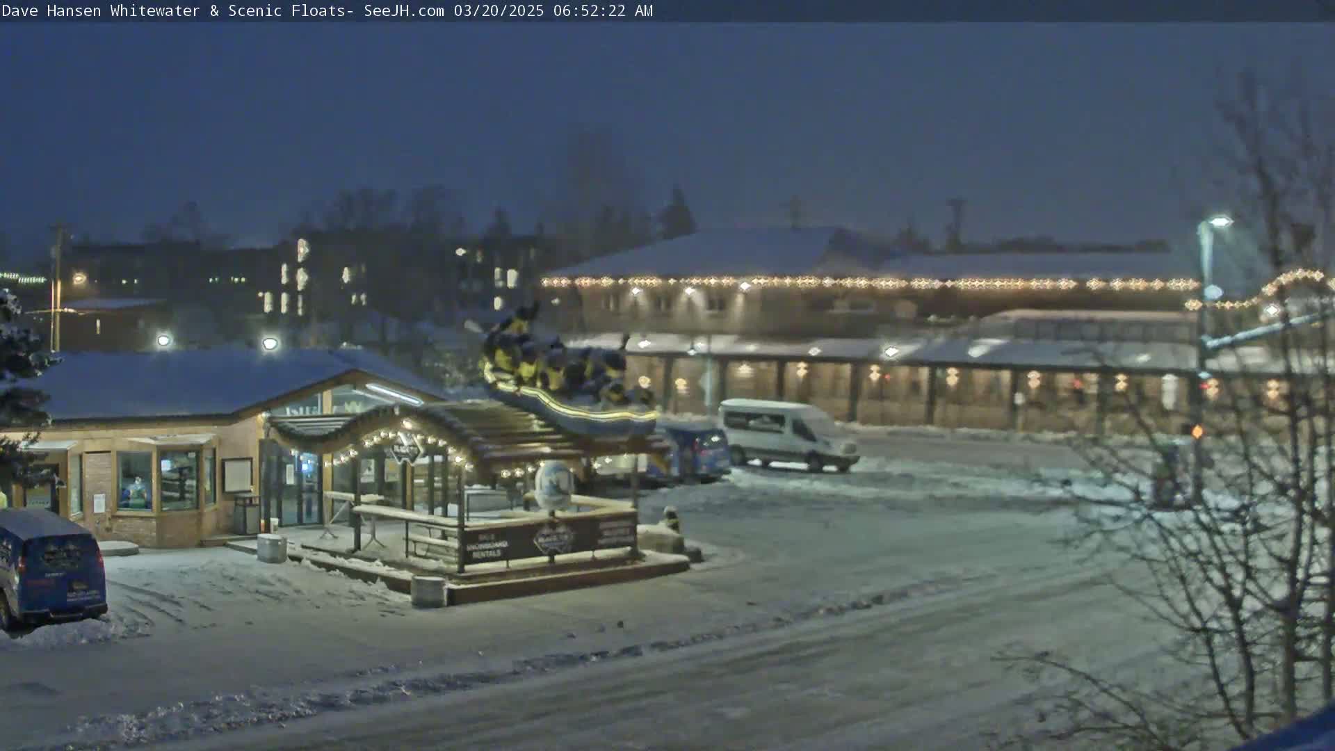 A snow-covered nighttime scene shows a building with string lights, a raft on a display stand, vehicles in a parking lot, and bare trees.
