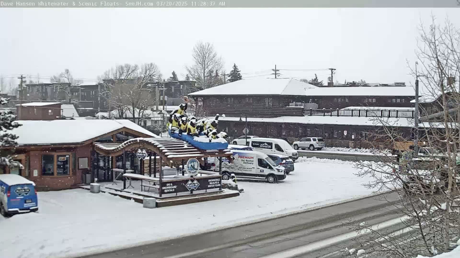 A snow-covered scene shows a building with a raft ride display in front, several vehicles in a parking lot, and light snowfall.